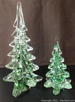 Photo showing two vintage glass Christmas trees, one taller (10h) and one smaller (6h), both clear with green accents on a black surface with brown background