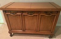 Front view of closed bar cabinet showing three paneled doors with brass handles, wood finish, and short legs on wheels.