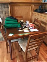 Wooden folding card table shown with four wooden folding chairs around it, four green chair covers folded on table. Various decks of playing cards and card game book on table surface.