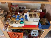 Photo shows shelf with assorted Christmas bulbs, some in boxes, and loose ornaments including blue, gold, silver and green colors, as well as a gold tinsel decoration.