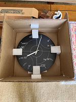Photo of new black clock with white mathematical notation in cardboard box with protective foam.