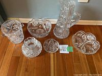 Top-down and angled view of six crystal glassware items including a tall pitcher, ice bucket, vase, two bowls, and a small star-shaped dish placed on wooden floor against wall.