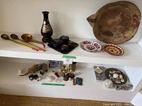 Shelf displaying a variety of decorative items including wooden spoons, vase, carved figurines, brass candle holders, stones, and a large wooden bowl with two decorative plates below.