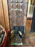Brass fireplace tool set with poker, brush, and shovel on a stand, placed near fireplace tiles alongside a basket with firewood logs.
