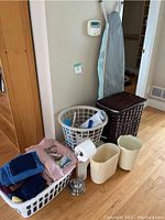 First photo showing plastic laundry baskets, folded clothing, toiletries, wicker hamper, ironing board, two white scalloped wastebaskets, and small metal toilet paper holder.