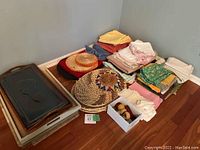 Overall view of assorted linens folded and stacked, napkin rings in a box, hats, and woven basket/tray on hardwood floor by wall.