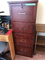 Photo of four-drawer dark wooden filing cabinet with silver handles and lock on top drawer, placed beside desk and side stand with wheels.