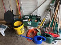 Photo showing assortment of garden and cleaning tools leaning against wall including watering cans, shovels, buckets, and plant stands.