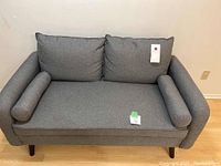 Front view of small grey sofa showing back cushions and bolster pillows, overall condition visible with fabric upholstery and dark tapered legs.