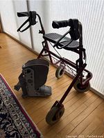 Red metal walker with black handles, brakes, seat, and wire basket, paired with grey air cast boot