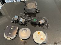 Photo showing all three portable CD players, their power adapters, and the carrying case on a black surface.