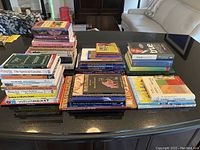 Top view of three main stacks of books on a black surface with a sofa and chairs in the background. Books cover topics including art, history, travel, ancient Egypt, interior design, and financial planning.