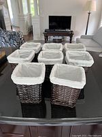 Eight square wicker baskets with white fabric liners displayed on a black table in a living room setting.