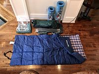 Photo showing assembled sleeping bag, two Airolite sleeping pads rolled upright, green tent fabric, tent poles, and tent pegs laid on wooden floor