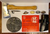 Photo showing the fire brigade axe with bright polished nickel head and wooden handle, three achievement badges in gold, silver and bronze, red commemorative book, black leather axe case, and a large circular anniversary plaque.