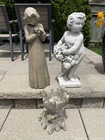 Front view of three cement garden statues showing the Guardian Angel holding a cat, Bacchus holding grapes, and the Tree Beard planter resembling a tree stump with carved faces.