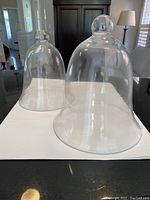 Two clear glass cloches on a white surface, large on right and medium on left, showing bell-shaped domes with knob handle tops.