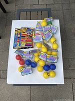 Photo showing all items in lot on white surface: 3 packs of colored wooden PopSticks and 17 packs of Happy Face Squeeze Balls in assorted colors.