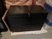Front view of the matte black metal filing cabinet with two drawers closed, placed over a white cloth on the floor.