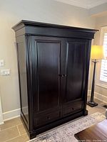 Solid wood armoire front view showing two paneled doors and two lower drawers in dark espresso color with crown molding and column details.