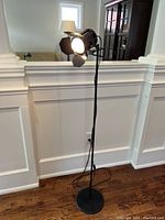 Full view of the matte black metal floor lamp standing on hardwood floor against white paneled wall, showing the lamp at full height with round base and power cord.