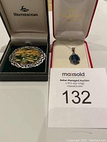 Image showing two pieces of jewelry in boxes: a brooch with a large patterned oval stone in a woven silver-tone frame, and a necklace with a blue-green opalescent pendant on a sterling silver chain.
