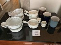 Overview of the lot showing two white casserole dishes with lids, four mugs in different styles, and two small ceramic cups arranged on a black surface.