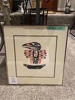 Framed Indigenous art print with eagle-like bird motif in black and red. Signed by artist in lower right corner. Dimensions 20x19 inches visible on tape above frame. Photo shows front view with some glare on glass.