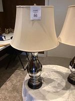 Single silver-coloured table lamp with white fabric shade on a table. Label with lot number attached to shade.
