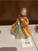 Front view of Royal Doulton ceramic figurine 'Rachel' showing figure wearing an orange hooded coat with fur edges and a green dress. Figurine is missing one foot.