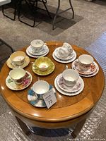 Overall photo showing 7 tea cups and matching saucers in various floral and decorative patterns on round wooden table.