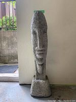 Front view of the tall, textured concrete sculpture depicting a stylized human face on a square base.