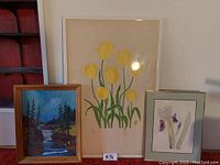 Three framed art pieces leaning against a wall, showing the group with the floral print of yellow tulips central and biggest, the purple iris floral print on the right, and the landscape painting on the left.