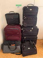 Six pieces of luggage stacked against wall including black and maroon suitcases and bags