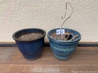 Two blue glazed ceramic garden pots side by side on a wooden deck against a stucco wall; one dark blue and one mixed blue-green with soil and dried plant stems