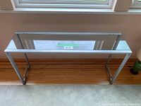 Frontal view of the clear glass top console table standing next to a window, showing its length and clear surface.