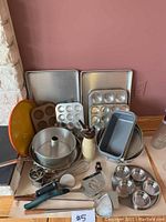Kitchen and bakeware items arranged together showing cookie sheets, muffin tins of different sizes, multiple cake pans, a stainless steel frying pan, colander, and an assortment of kitchen tools including a slicer and peeler.