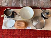 Overview of entire kitchenware lot showing fondue pot with burner and forks, large mixing bowl, covered pan with lid, and several ceramic serving dishes on a red carpet background.