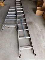 Two large aluminum extension ladders lying flat on a concrete floor, showing full length and detailing of rungs, locking mechanisms, and blue rope on one ladder.