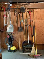 Photo showing Karcher electric pressure washer on floor with hose and cord, multiple garden tools including shovels, rakes, broom, manual lawn mower mounted on wall