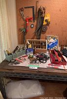 View of hand tools, tape measures, caulking gun, organizer tray with hardware and tools, hanging tools including clamp, hand saws, and measuring square above a metal workbench with a plastic drop sheet underneath.