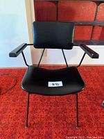 Front view of chair showing black seat, backrest, wooden arms, and metal legs on red patterned carpet