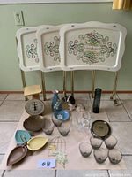 Full lot overview showing three vintage TV trays, antique food scale, assorted glassware, brass plate, vases, dog figurine, leaf-shaped dishes, and decorative items arranged on tiled floor.
