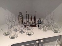 Wide shot of complete barware set with glasses, cocktail bowls, shaker, and tool set on white counter.