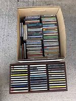 Full view of a cardboard box filled with about 60 assorted music CDs on a concrete surface