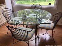View of entire round glass top dining table with four metal frame chairs showing seat cushion pattern and metal frames.