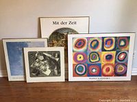 Four framed art prints leaning against a wall on floor. Central item is colorful Kandinsky print with circular motifs. Other prints are smaller with black and white or muted tones.