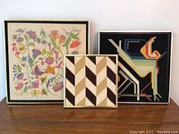 Three framed embroidery/needlepoint artworks displayed on a wooden surface against a white wall, showing an overall view of the lot