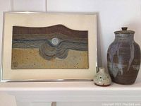 Framed art with abstract design and two pottery vases on a white shelf, side angle