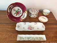 Overview of all porcelain items in the lot arranged on a wooden surface: burgundy and gold-trimmed plate, floral trinket box, dishes with floral patterns.
